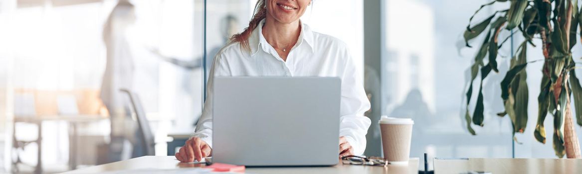 Portrait of smiling lady boss working on laptop at workplace in modern office. Blurred background