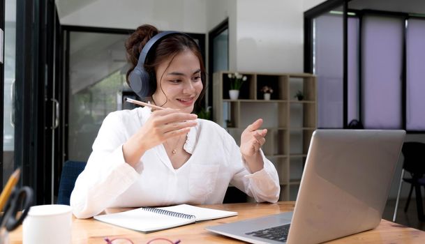 Smiling young asian woman in headset wave greet talking on webcam virtual conversation on laptop, happy female in wireless headphones speak on video call on computer, consult client online.