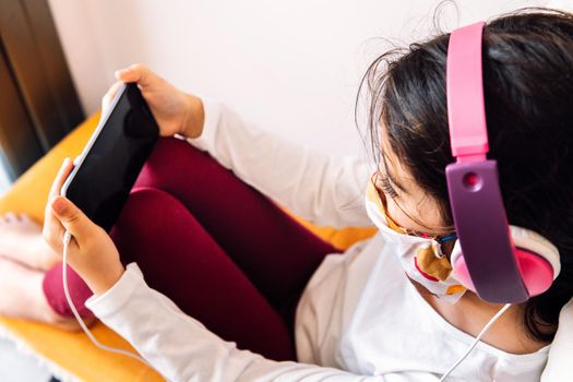 little girl sitting at home with a headset and protective mask learning with the phone, coronavirus concept and home entertainment for children