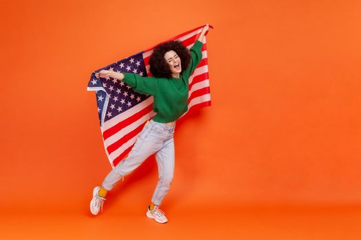 Full length photo of woman with Afro hairstyle wearing green casual style sweater raised arms, holding american flag, celebrating national holiday. Indoor studio shot isolated on orange background.