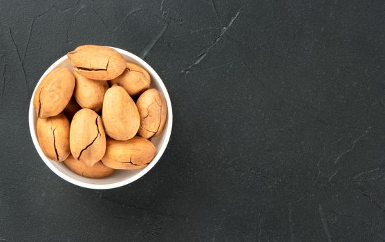 Full bowl of dry inshell pecans on empty dark background, top view.