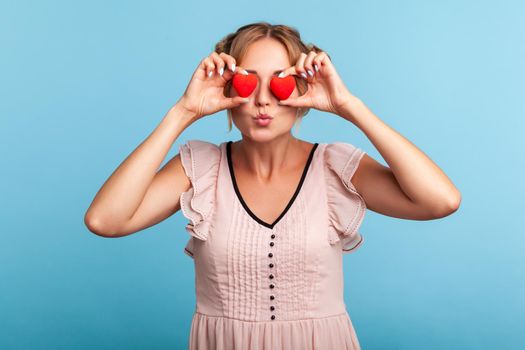 Portrait of lovely charming blonde female with hair buns holding red hearts and sending air kiss, sharing love, expressing amorous feelings. Indoor studio shot isolated on blue background.