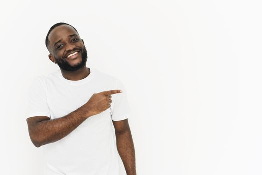 Smiling african american man in white t-shirt pointing with finger to the side at copy space for your text ad promoting your product in white background.