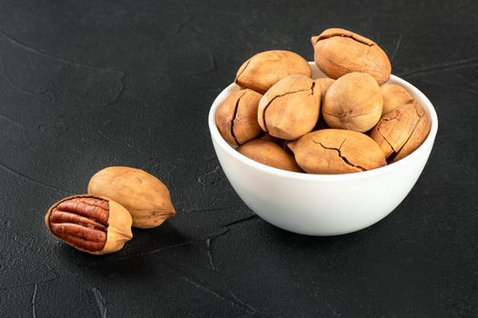 Two dry pecans with a full white bowl of nuts close-up on a dark background.