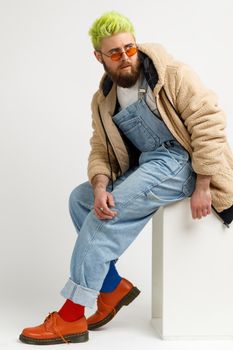 Young man with beard and trendy green hair wearing stylish clothing and shoes, sitting on white cube and looking away with pensive expression. Indoor studio shot isolated over gray background.
