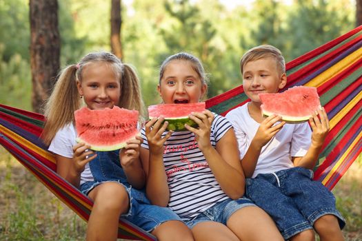 three cheerful children eat watermelon and joke, outdoor, sitting on a colorful hammock. Summer fun and leisure