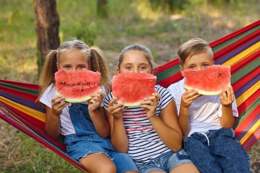 three cheerful children eat watermelon and joke, outdoor, sitting on a colorful hammock. Summer fun and leisure