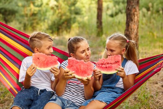 three cheerful children eat watermelon and joke, outdoor, sitting on a colorful hammock. Summer fun and leisure