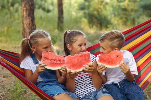 three cheerful children eat watermelon and joke, outdoor, sitting on a colorful hammock. Summer fun and leisure