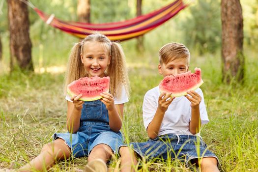 Happy Kids eating watermelon in the park. Kids eat fruit outdoors. Healthy snack for children. Little girl and boy playing in the forest biting a slice of water melon