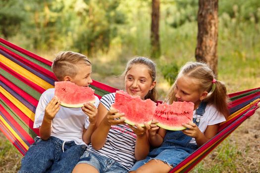 three cheerful children eat watermelon and joke, outdoor, sitting on a colorful hammock. Summer fun and leisure