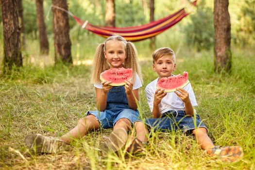 Kids eating watermelon in the park. Kids eat fruit outdoors. Healthy snack for children. Little girl and boy playing in the forest biting a slice of water melon