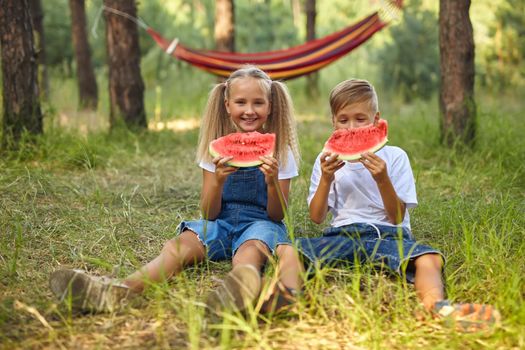 Kids eating watermelon in the park. Kids eat fruit outdoors. Healthy snack for children. Little girl and boy playing in the forest biting a slice of water melon