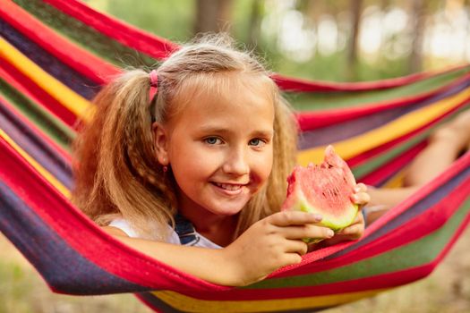 Cute little girl resting in a colored hammock in the forest and eating fresh watermelon