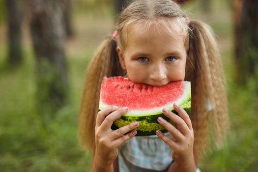 Close up portrait of Funny girl eating watermelon in the park. Kid eat fruit outdoors. Healthy snack for children.