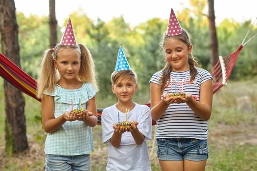 Children holding birthday cakes with burning candles. Kids party decoration and food. Boy and girls celebrating birthday in the garden with hammock. Kids with sweets.