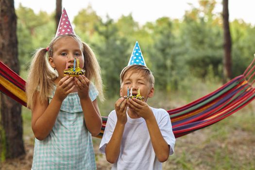 Children eating birthday cakes with burning candles. Kids party decoration and food. Boy and girl celebrating birthday in the garden with hammock. Kids with sweets.