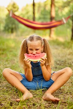 Funny girl eating watermelon in the park. Kid eat fruit outdoors. Healthy snack for children. Little girl playing in the forest biting a slice of water melon