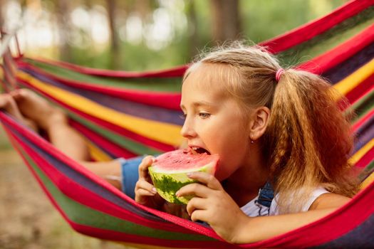 Cute little girl resting in a colored hammock in the forest and eating fresh watermelon