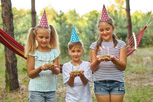 Children holding birthday cakes with burning candles. Kids party decoration and food. Boy and girls celebrating birthday in the garden with hammock. Kids with sweets.