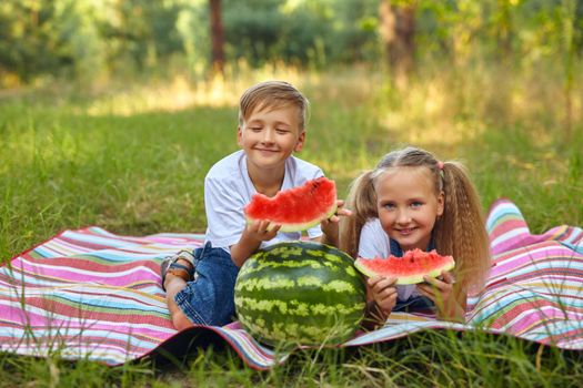 Kids eating watermelon in the park. Kids eat fruit outdoors. Healthy snack for children. Little girl and boy playing in the forest biting a slice of water melon