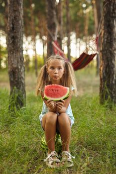 Funny girl eating watermelon in the park. Kid sitting on a big watermelon outdoors. Healthy snack for children. Little girl playing in the forest biting a slice of water melon