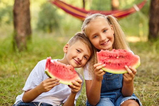 Kids eating watermelon in the park. Kids eat fruit outdoors. Healthy snack for children. Little girl and boy playing in the forest biting a slice of water melon