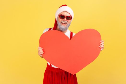 Positive cheerful elderly man in trendy sunglasses and santa claus hat holding and showing paper heart, symbol of love, winter holidays. Indoor studio shot isolated on yellow background