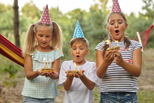 Children blow candles on birthday cake. Kids party decoration and food. Boy and girls celebrating birthday in the garden with hammock. Kids with sweets.