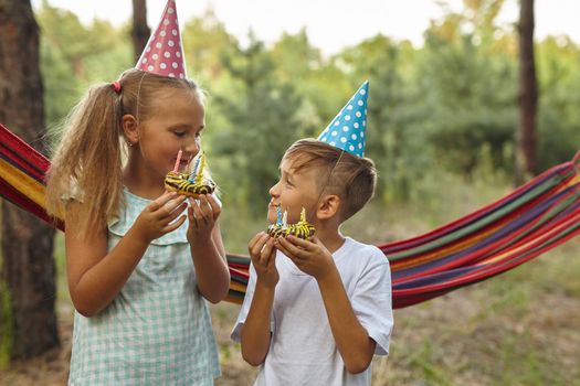 Children eating birthday cakes with burning candles. Kids party decoration and food. Boy and girl celebrating birthday in the garden with hammock. Kids with sweets.