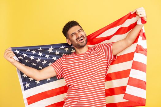 Excited patriotic man with beard in striped t-shirt standing holding in hands flag of united states of america and screaming, singing anthem. Indoor studio shot isolated on yellow background