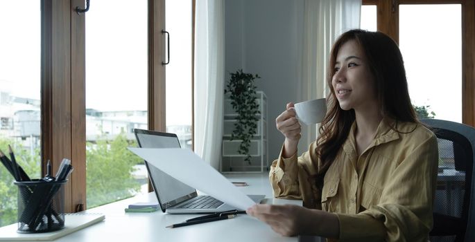 Beautiful young Asian businesswoman drinking a coffee working on laptop at office..