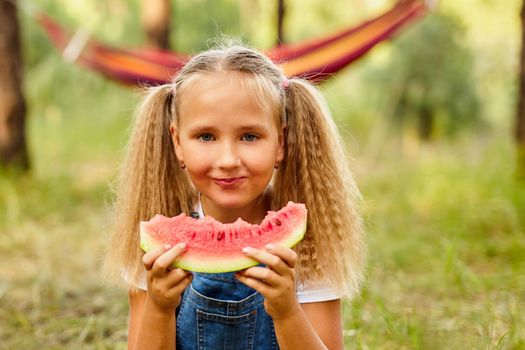 Funny girl eating watermelon in the park. Kid eat fruit outdoors. Healthy snack for children. Little girl playing in the forest biting a slice of water melon