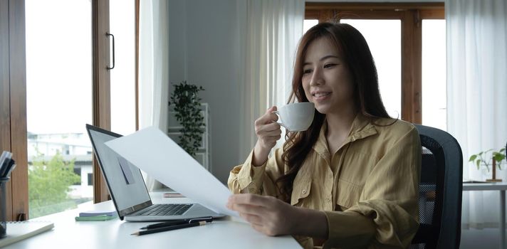 Beautiful young Asian businesswoman drinking a coffee working on laptop at office..