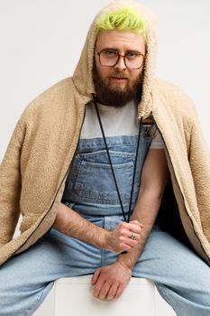 Attractive young adult model man sitting in white cube and looking at camera, wearing denim overalls and jacket with hood, with calm expression. Indoor studio shot isolated over gray background.