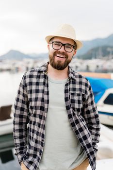Handsome man wearing hat and glasses near marina with yachts. Portrait laughing man with sea port background.
