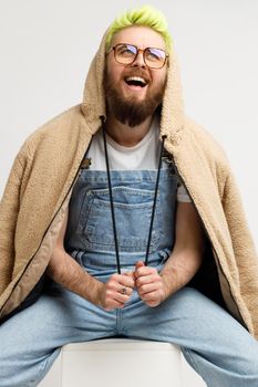 Bearded man wearing fashionable jacket and denim overalls, expressing positive emotions, laughing, looking up with optimism, sitting on cube. Indoor studio shot isolated over gray background.