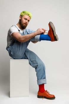 Bearded hipster man sitting on white cube. raising his leg and ties his shoelaces, looking at camera, wearing trendy denim overalls, has serious look. Indoor studio shot isolated over gray background.