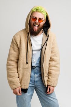 Handsome bearded man wears in stylish jacket and denim overalls fashion hair style looking at camera with crazy expression, showing tongue. Indoor studio shot isolated over gray background.