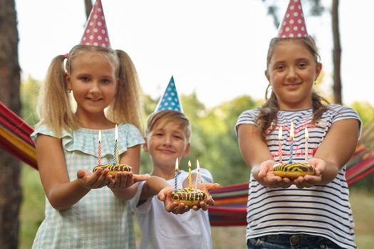 Children holding birthday cakes with burning candles. Kids party decoration and food. Boy and girls celebrating birthday in the garden with hammock. Kids with sweets.