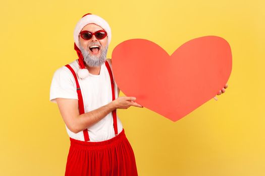 Happy cheerful man in holiday costume, santa claus hat and stylish sunglasses holding big red paper heart, symbol of affection, saint valentine day. Indoor studio shot isolated on yellow background