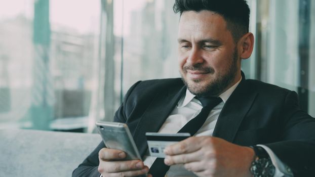 Portrait of cheery bearded Caucasian businessman in formal clothes smiling and paying online bill while keeping credit card and smartphone in his hands in glassy cafe during lunch break.