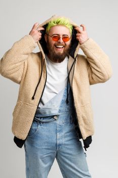 Happy hipster model with beard and bright hair, looking at camera and laughing loud out, putting on hood, expressing positive emotions. Indoor studio shot isolated over gray background.