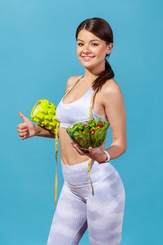Full length profile portrait satisfied woman in white sportswear with tape measure showing thumbs up holding bowl with salad and roller massager, slimming. Studio shot isolated on blue background