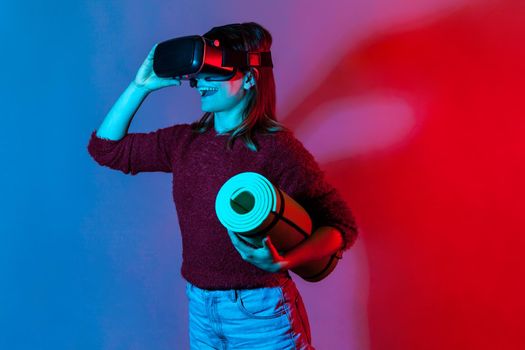 Happy girl with VR glasses headset holding rolled yoga mat, watching fitness courses and smiling excited about sports activity in virtual reality, workouts simulator. Innovation, technology concept
