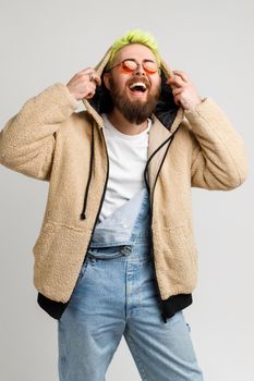 Extremely happy bearded young adult man wearing denim overalls and jacket, laughing out loud, keeping hands on his hood, hears funny joke. Indoor studio shot isolated over gray background.