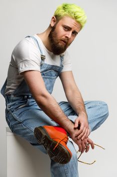 Young adult handsome guy wears fashionable denim overalls, white t shirt and red shoes, looking at camera, expressing sadness, holds glasses in hands. Indoor studio shot isolated over gray background.