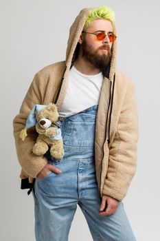 Attractive bearded man model wearing fashionable closing, has Teddy bear as accessory, keeping hand in pocket, looks away with thoughtful expression. Indoor studio shot isolated over gray background.