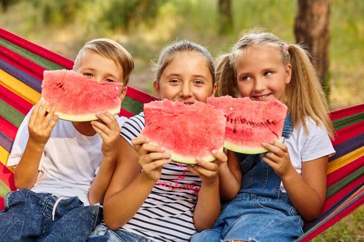 three cheerful children eat watermelon and joke, outdoor, sitting on a colorful hammock. Summer fun and leisure