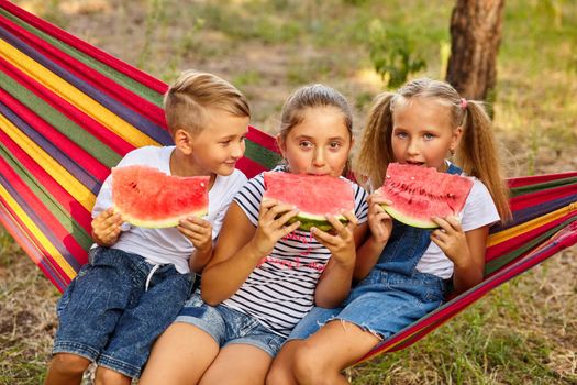three cheerful children eat watermelon and joke, outdoor, sitting on a colorful hammock. Summer fun and leisure
